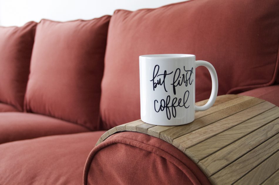 Close-up of a mug saying "But first, coffee" on top of a natural wood sofa armrest tray which rests on an Ektorp sofa in Comfort Works' custom red woollen slipcovers